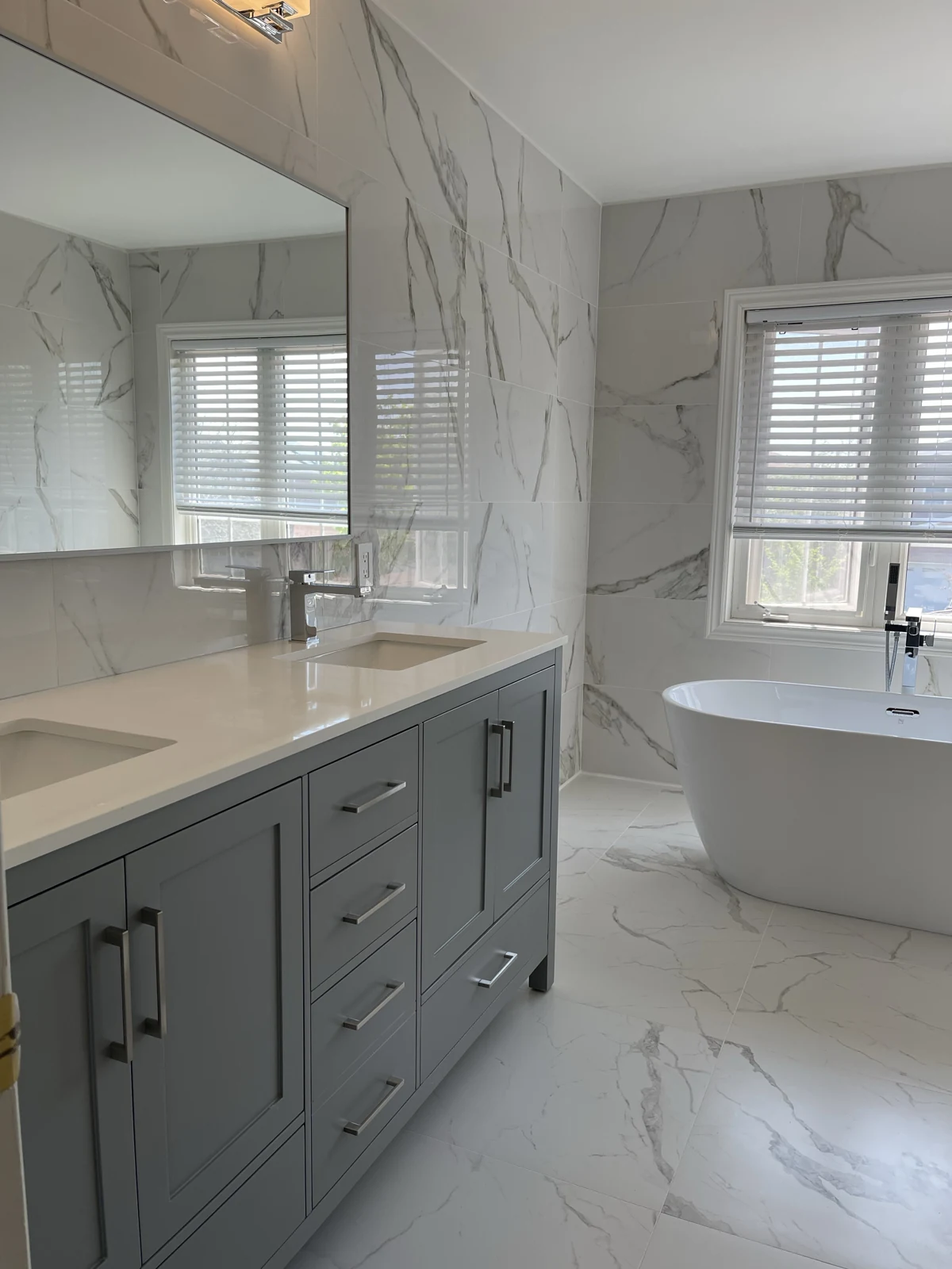 Grey double vanity and freestanding bath below a window, surrounded by pale marble-look wall and floor tile