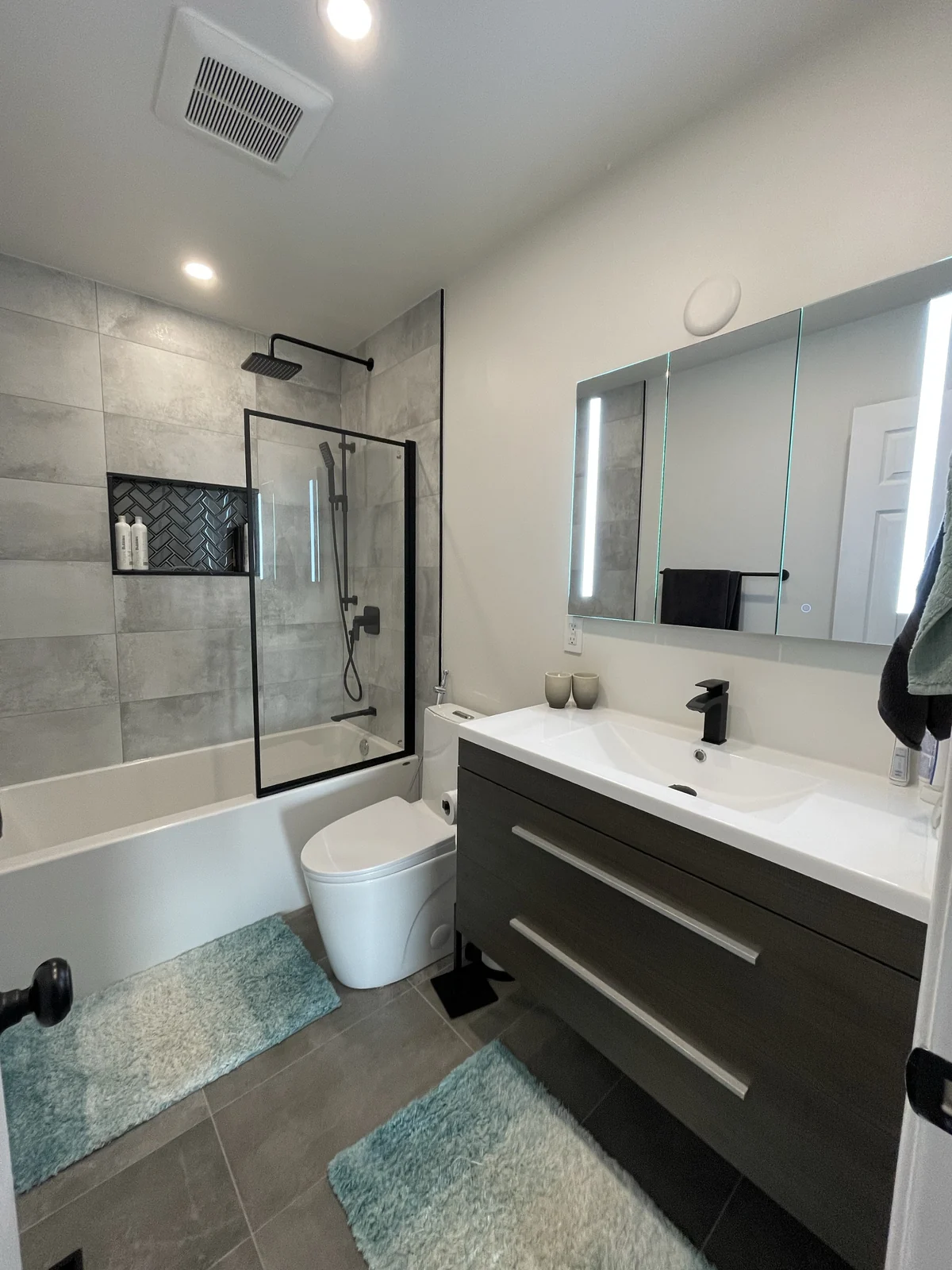 Bathroom with a floating wood-tone drawer vanity, illuminated mirror cabinet and built-in bath with a black-framed shower screen