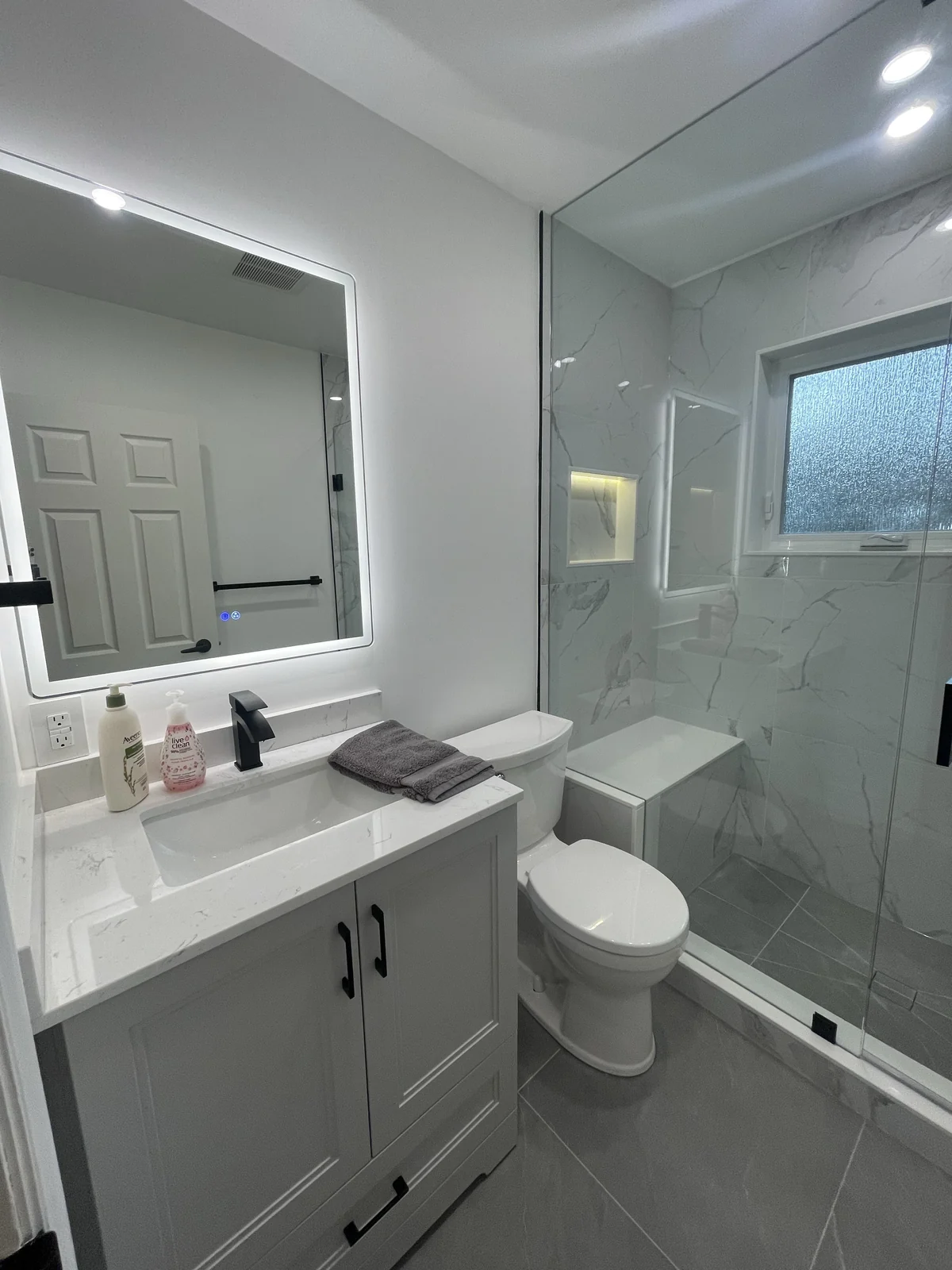 Light grey vanity and illuminated mirror beside a glass shower with a built-in bench, lit niche and frosted window