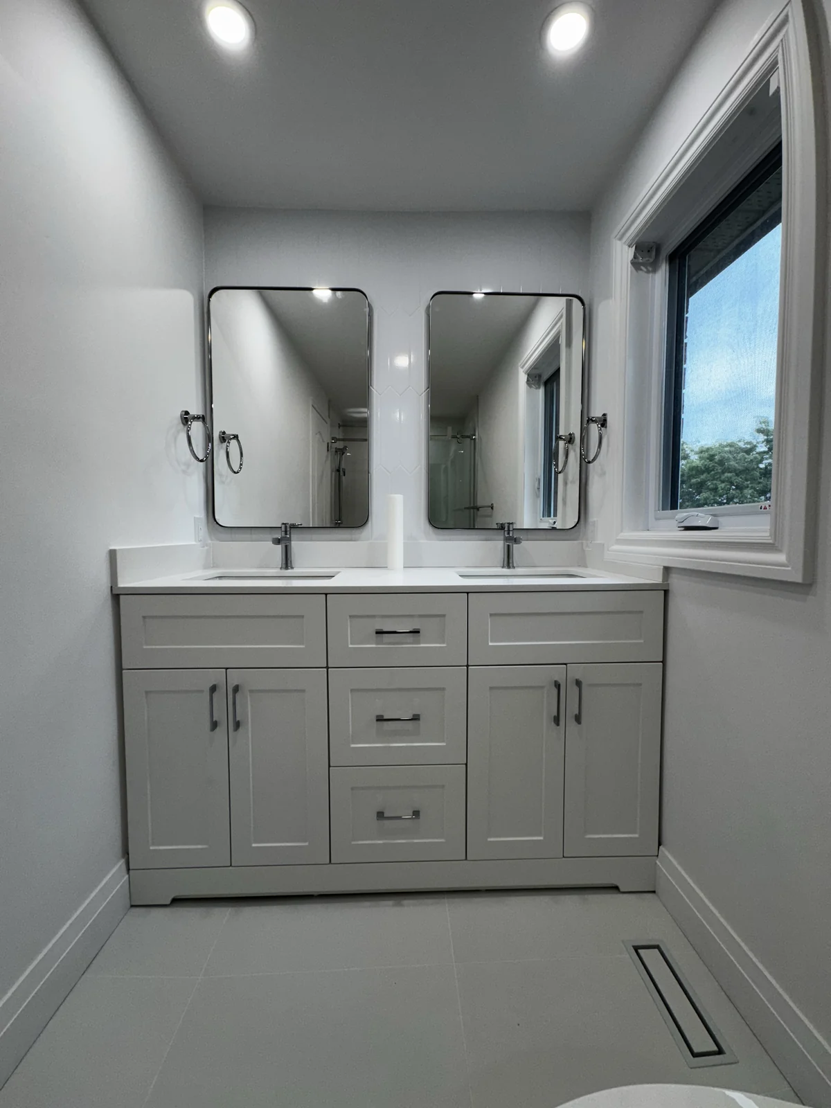 Light-coloured double vanity with central drawers, cupboard storage and two rounded-corner mirrors beside a window