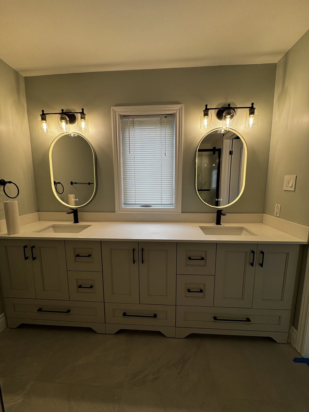 Double vanity with black handles and taps, two illuminated oval mirrors and glass-shaded lights flanking a central window