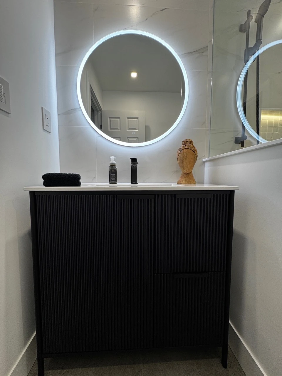 Black fluted vanity with drawer and cupboard storage, a round illuminated mirror and marble-look wall tile