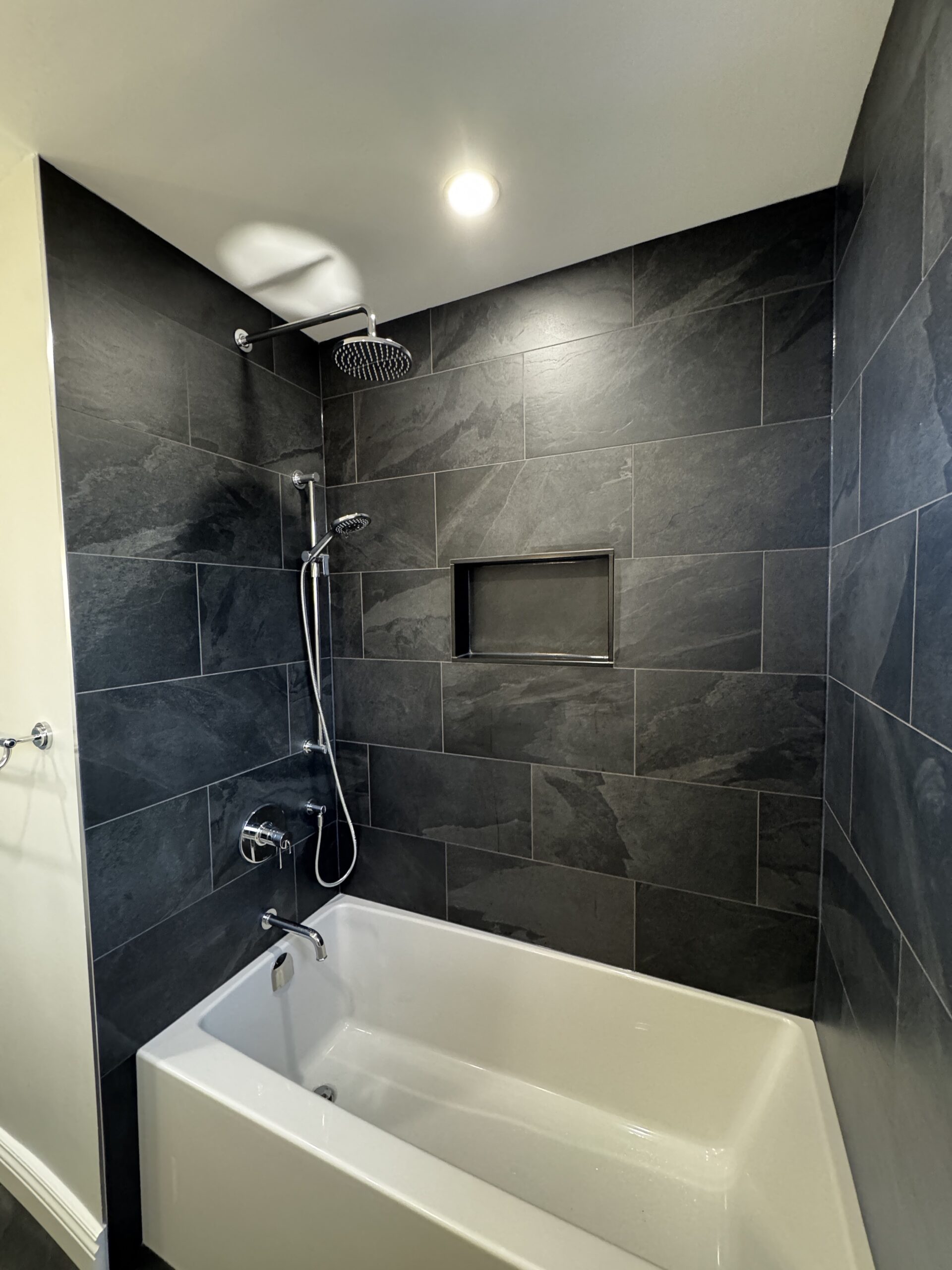 After bathroom renovation in Clarkson, Mississauga: a white bathtub with dark rectangular wall tiles, a recessed niche, an overhead shower and a handheld shower.