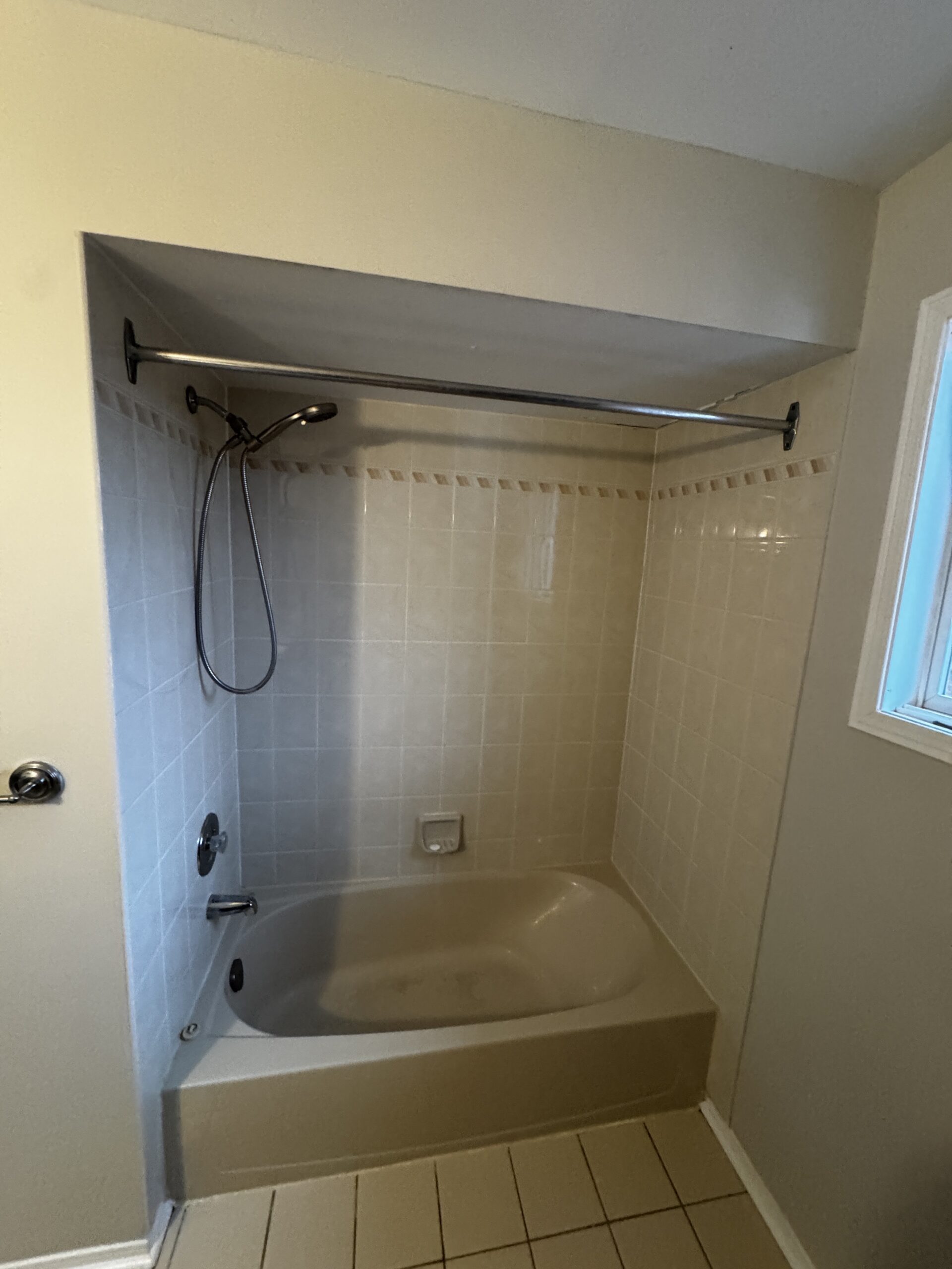 Before bathroom renovation in Clarkson, Mississauga: a bathtub with light square wall tiles, a decorative border, a curtain rod and a handheld shower.