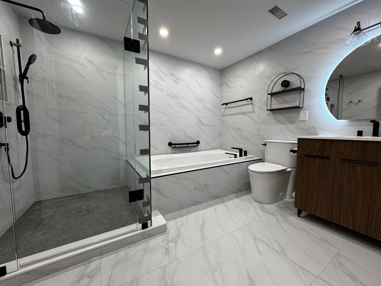 Finished Erin Mills bathroom showing a glass shower, separate bathtub, wood-tone vanity, illuminated mirror and white marble-look wall and floor tiles.