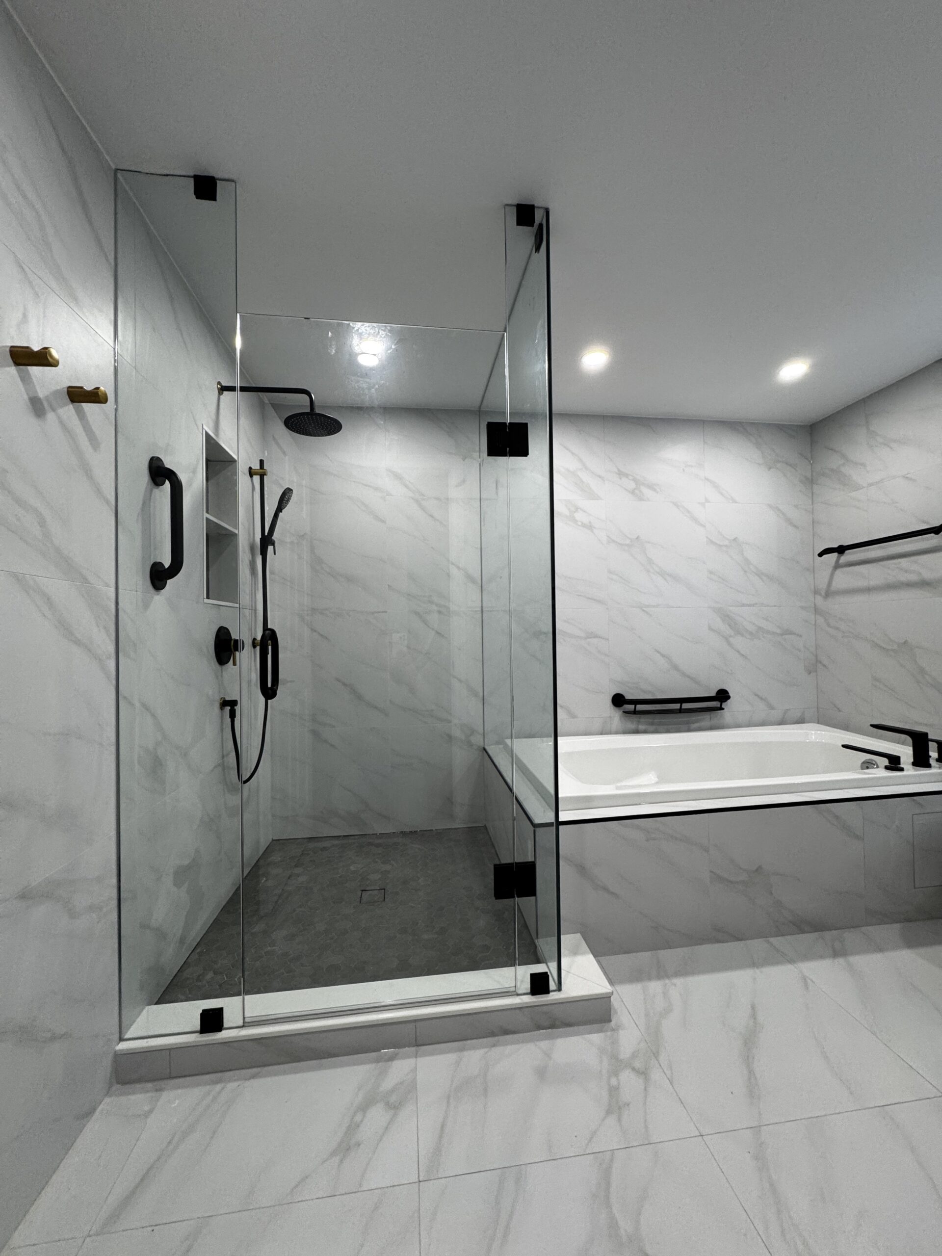 Renovated glass shower in Erin Mills with black fixtures, recessed shelving, grey small-format floor tile and a raised threshold beside the bathtub.