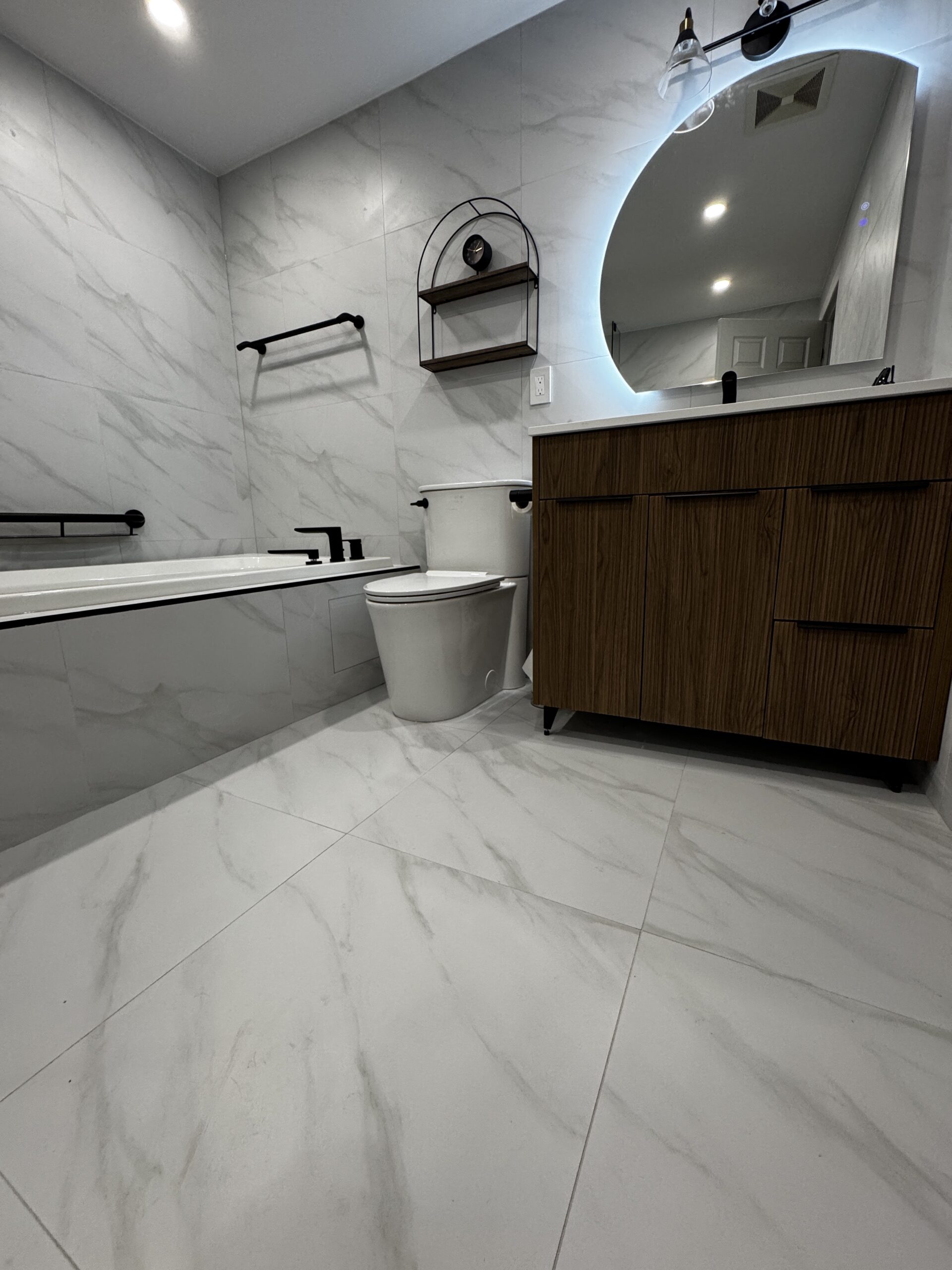 After renovation in Erin Mills: a wood-tone vanity, rounded illuminated mirror, black fixtures and white marble-look tile around the bathtub.