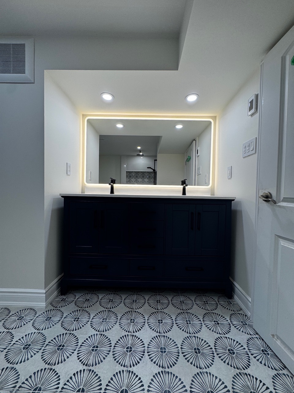 Dark double vanity beneath a large illuminated mirror with patterned flooring in Erin Mills