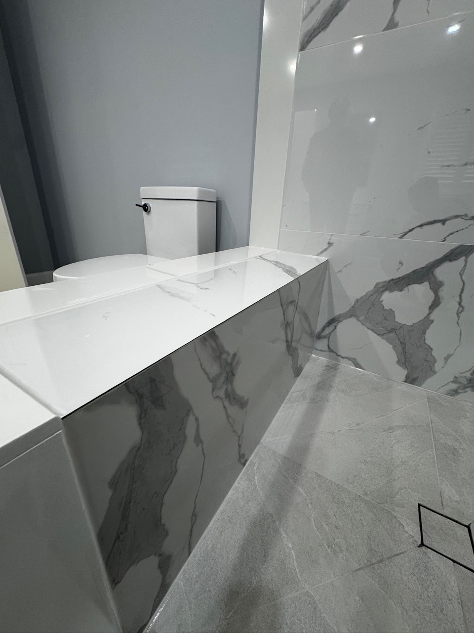 Marble-look shower bench beside grey floor tile and a square drain in Erin Mills