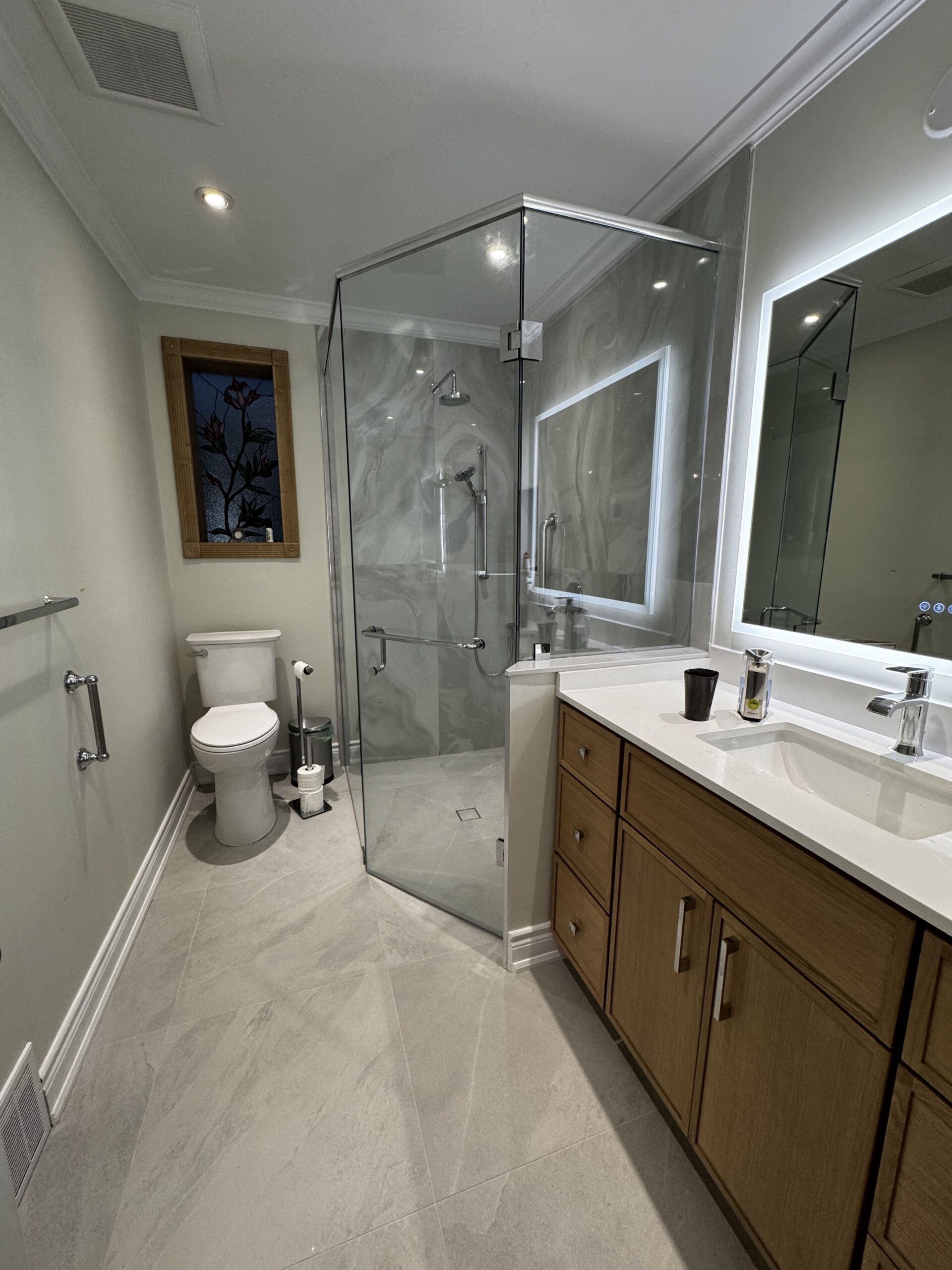 After renovation in Mineola, Mississauga: a curbless glass shower with grey veined tiles, a wood-tone vanity, an illuminated mirror and the stained-glass window.