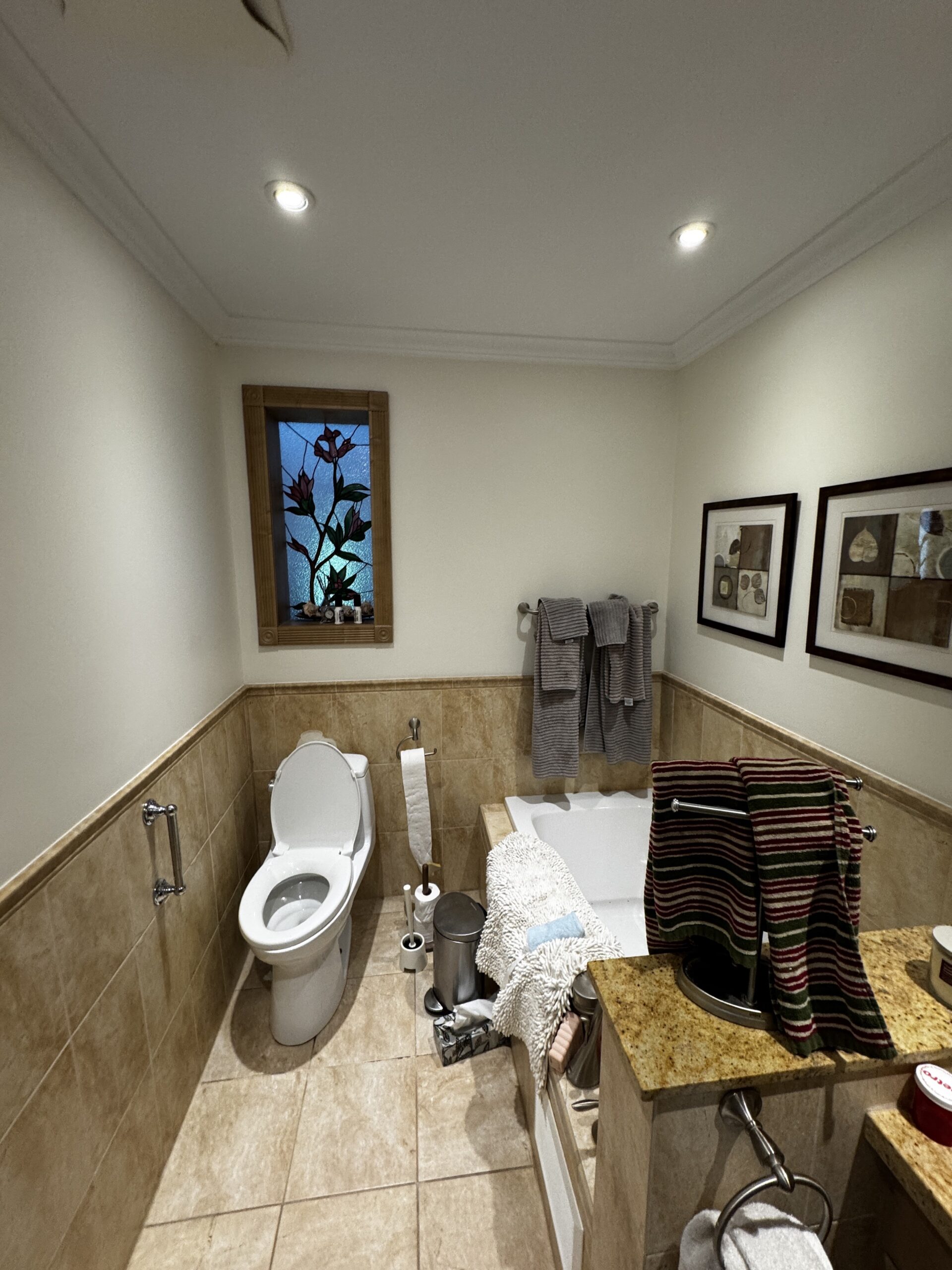 Before bathroom renovation in Mineola, Mississauga: a bathtub beside the toilet, beige wall and floor tiles, and a floral stained-glass window.