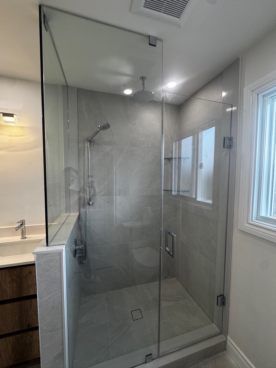 After renovation: clear glass shower with large grey tiles, an overhead shower, a handheld shower, glass shelves and a wood-tone vanity beside it.