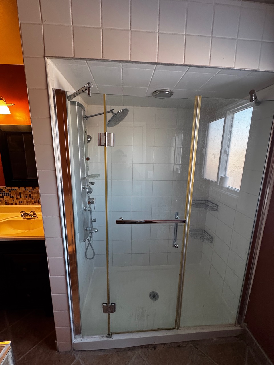 Before renovation: shower with white square tiles, metal-trimmed glass doors and a shower panel beside a frosted window.